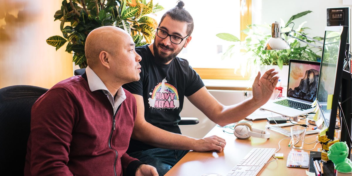 Die Softwareentwickler Tung Ngo und Niko Gomes von Micromata im Gespräch an Nikos Schreibtisch. Im Hintergrund eine Grünpflanze