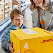 Mutter und Sohn reichen ein DHL-Paket über den Tresen eines DHL-Paketshops. Motiv aus der offiziellen DHL-Mediathek.