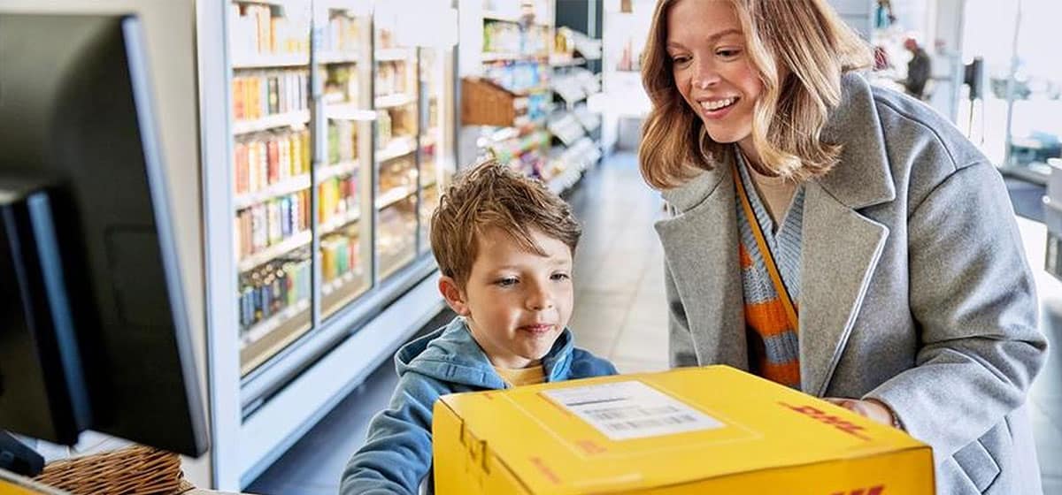Mutter und Sohn reichen ein DHL-Paket über den Tresen eines DHL-Paketshops. Motiv aus der offiziellen DHL-Mediathek.